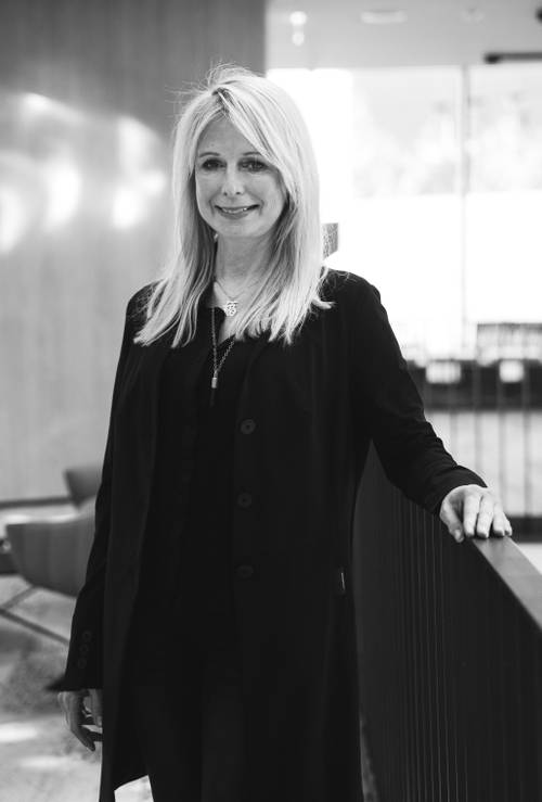 Schwarz-Weiß-Foto einer Frau mit langen blonden Haaren, die einen dunklen Mantel trägt und sich in einer modernen Innenausstattung an ein Geländer lehnt.