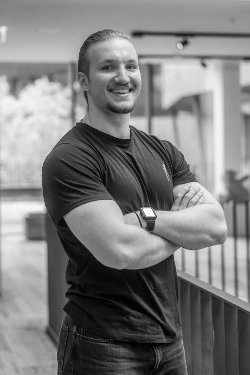 Lächelnde Person mit langen Haaren, schwarzem T-Shirt und Smartwatch, die mit verschränkten Armen in einem gut beleuchteten Raum steht.