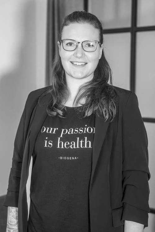 Eine Person mit Brille, lächelnd, mit einem T-Shirt mit der Aufschrift „Our passion is health” und einem Blazer, stehend in einem Innenraum.