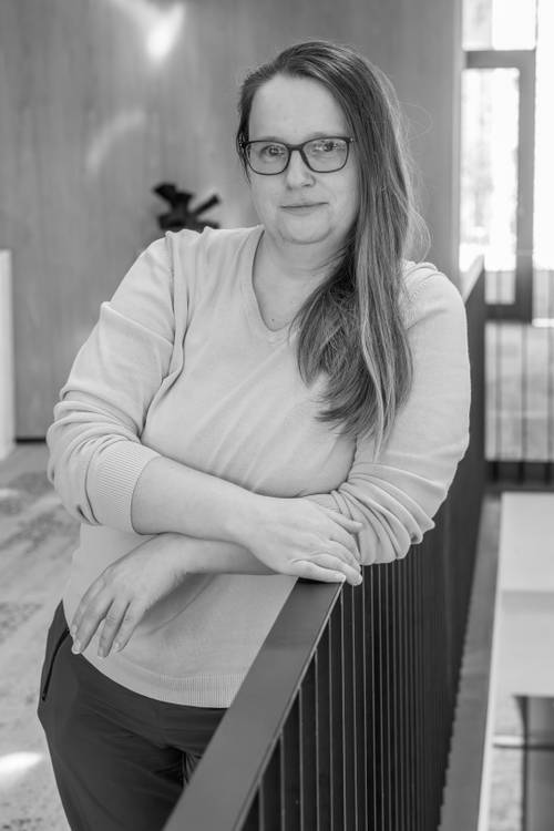 Frau mit Brille lehnt sich in einem modernen Interieur an ein Geländer, trägt einen leichten Pullover und hat langes Haar über der Schulter. Schwarz-Weiß-Bild.