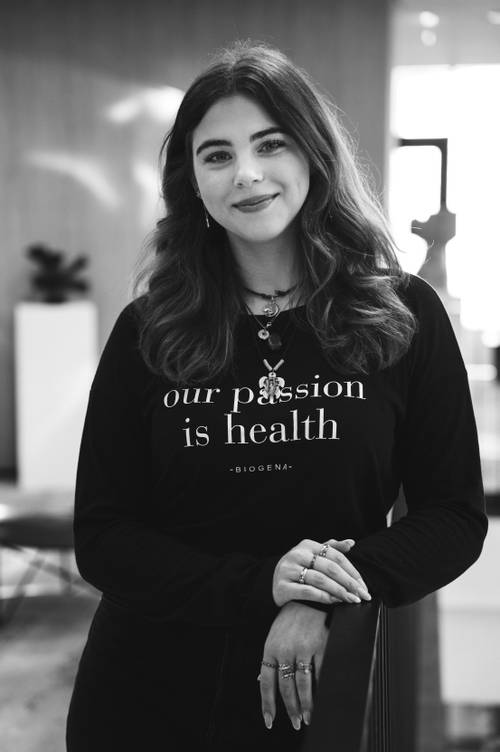 Eine Frau mit langen Haaren lächelt und trägt ein Shirt mit der Aufschrift „Our passion is health“. Sie steht in einem Innenraum und lehnt sich an ein Geländer.
