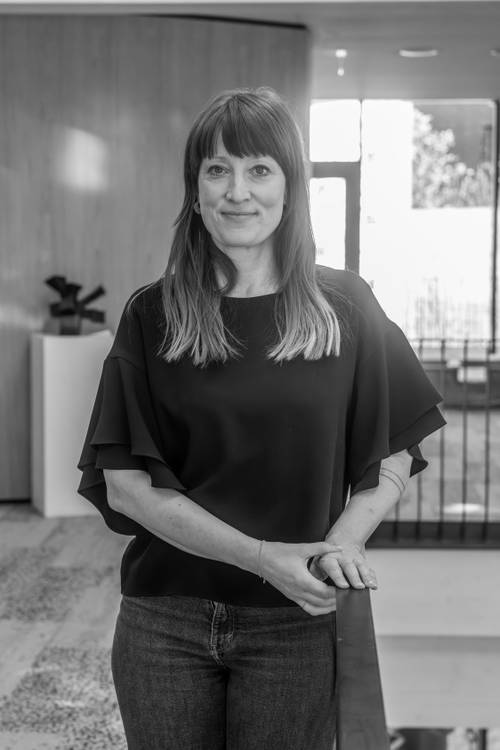 Eine Person mit langen Haaren, die eine dunkle Bluse und Jeans trägt und in einem modernen Innenraum mit Holz- und Glaselementen steht. Schwarz-Weiß-Foto.