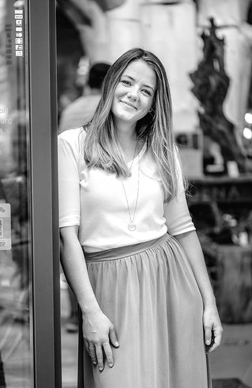 Lächelnde Frau mit langen Haaren, bekleidet mit einem weißen Oberteil und einem langen Rock, lehnt an einer Glastür in einer Stadtstraße. Schwarz-Weiß-Foto.