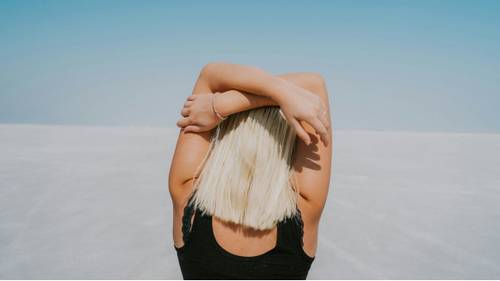 Person mit blonden Haaren, die ihre Arme über den Kopf streckt und auf einer weiten, weißen Salzfläche unter einem klaren blauen Himmel steht.
