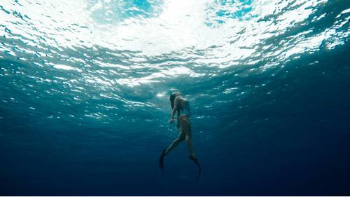 Eine Person in Badeanzug und Flossen schwimmt unter Wasser, umgeben vom tiefblauen Ozean und beleuchtet vom Sonnenlicht, das durch die Oberfläche scheint.
