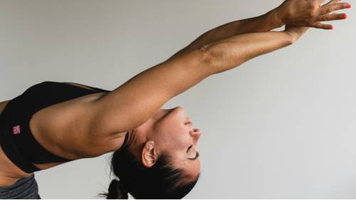 A person in athletic wear stretches with arms extended overhead, demonstrating a yoga pose against a plain background.