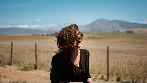 Eine Frau mit vom Wind zerzausten Haaren lacht fröhlich in einem weiten, offenen Feld mit Bergen im Hintergrund unter einem klaren blauen Himmel.