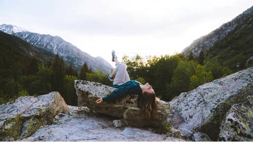 Person, die auf einem großen Felsen liegt, umgeben von Bergen und Bäumen, mit einem heiteren Himmel im Hintergrund.