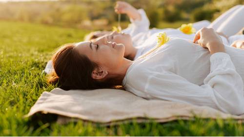 Two people in white shirts lying on a blanket in a grassy field, enjoying a sunny day with flowers nearby.