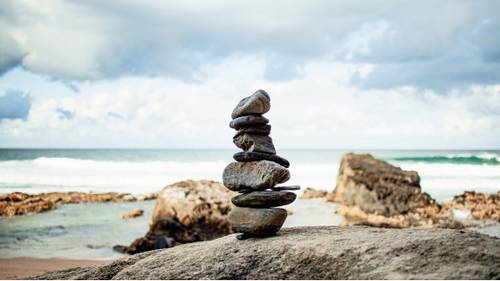 Stacked stones on a rocky beach with waves and a cloudy sky in the background.