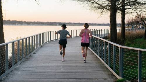 Two people jogging on a lakeside boardwalk at sunset, surrounded by trees and calm water.
