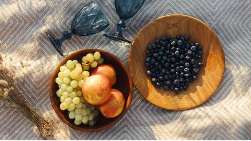 Two bowls on a picnic blanket: one with grapes and apples, the other with blueberries. Two blue glasses lie beside them.