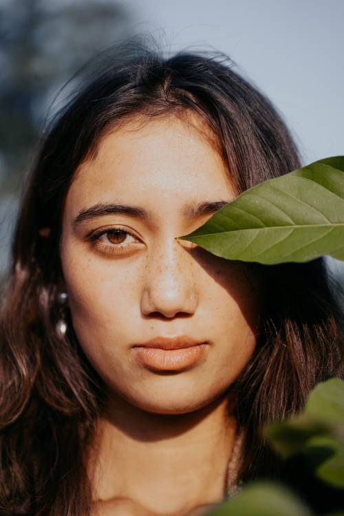 Prim-plan al unei femei cu părul lung, al cărei ochi este parțial acoperit de o frunză verde, în lumină naturală.