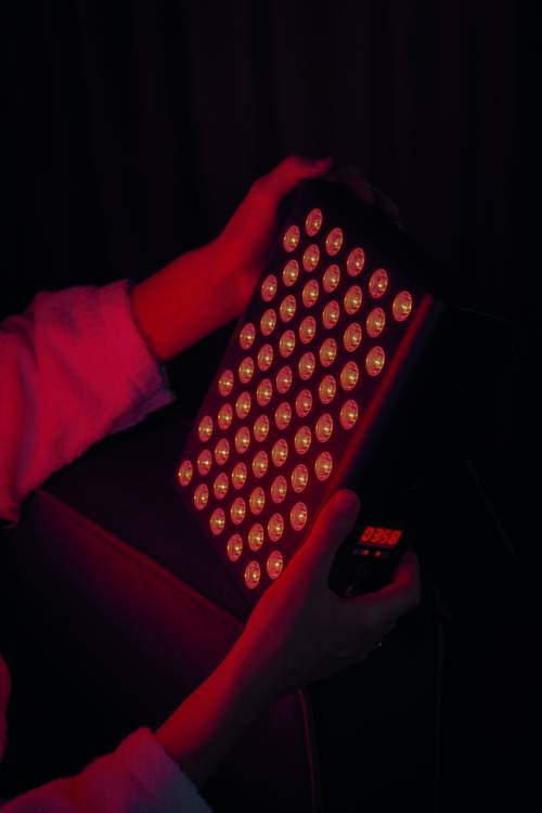 Hände, die ein rotes LED-Lichttherapie-Panel mit mehreren leuchtenden Lampen in einer dunklen Umgebung halten.