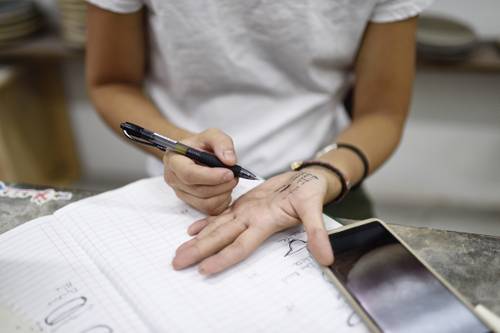 A person writing notes on their palm with a pen while looking at a notebook and a smartphone on the table.