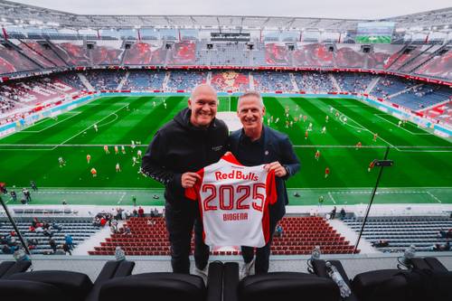 Zwei Männer halten ein Trikot mit der Aufschrift „Red Bulls 2025” in einem Stadion hoch, von dem aus man einen Blick auf einen Fußballplatz mit sich aufwärmenden Spielern hat.
