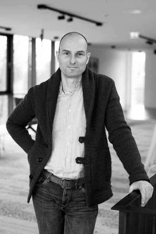 Mann mit dunklem Strickjacke und Jeans, der mit einer Hand auf einem Tisch in einem modernen, gut beleuchteten Raum steht. Schwarz-Weiß-Bild.