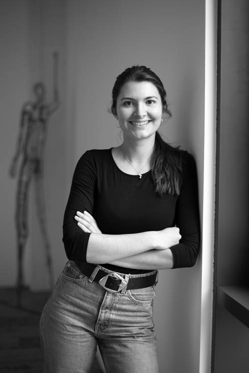 Eine Frau in einem schwarzen Oberteil und Jeans lehnt lächelnd mit verschränkten Armen an einer Wand. Im Hintergrund ist ein verschwommenes Skelettmodell zu sehen. Schwarz-weiß.
