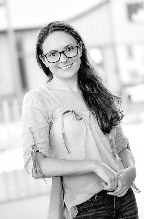Lächelnde Person mit langen Haaren und Brille, die eine helle Bluse trägt, steht im Freien vor einem unscharf dargestellten Gebäude.
