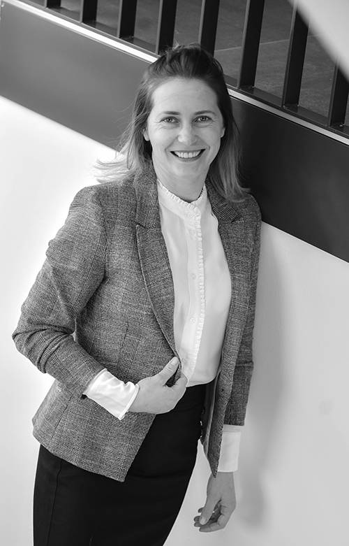 Lächelnde Frau in Blazer und Bluse, die drinnen an einer Wand mit Geländer steht. Schwarz-Weiß-Foto.