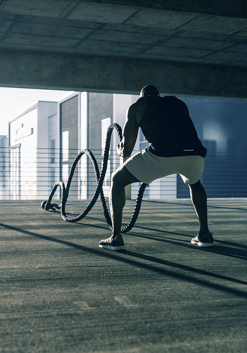Mann in Sportkleidung benutzt Seile auf einem Outdoor-Gym-Boden, mit einem modernen Gebäude im Hintergrund.