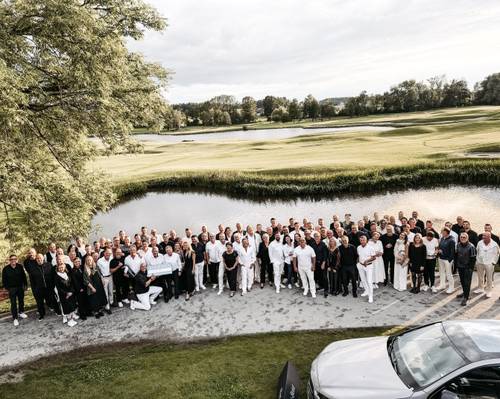 Eine große Gruppe von Menschen in weißer und schwarzer Kleidung versammelt sich im Freien in der Nähe eines Golfplatzes und eines Teiches, im Hintergrund sind Bäume und Gras zu sehen.
