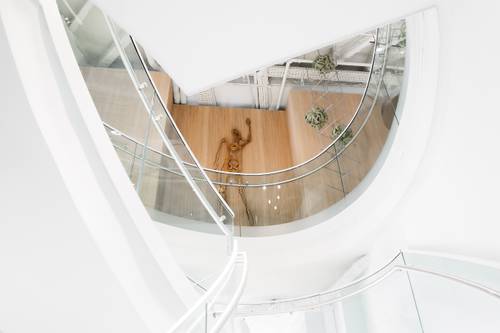 Blick von einer Wendeltreppe auf eine Holzfigur auf einem Holzboden, umgeben von klarer, moderner Architektur und Pflanzen.