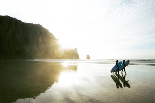 Two surfers with their boards are running along a sunny beach, with cliffs and the sea visible in the background.