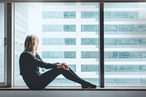 O femeie stă pe pervaz, privind afară spre un peisaj urban modern cu clădiri înalte, creând o atmosferă contemplativă.