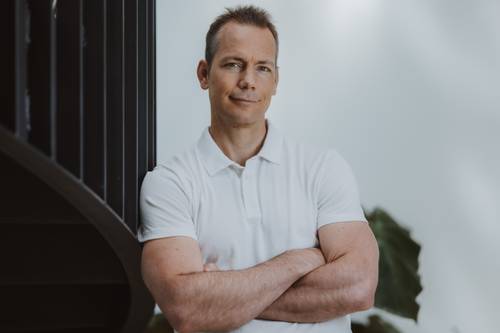 Ein Mann in einem weißen Poloshirt steht mit verschränkten Armen an einer dunklen Wendeltreppe, im Hintergrund ist eine Pflanze zu sehen.