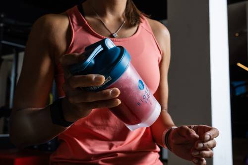 Eine Person in einem rosa Tanktop, die eine rote Shaker-Flasche mit blauem Deckel in einem Fitnessstudio in der Hand hält.