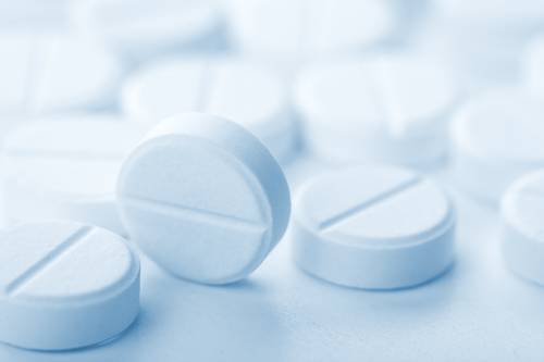 Nahaufnahme von mehreren weissen, runden Tabletten mit Bruchkerben, die auf einer hellen Oberfläche verstreut liegen, wobei eine Tablette scharf abgebildet ist.