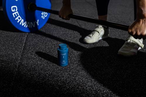 Person bereitet sich darauf vor, eine Langhantel mit blauen Gewichten zu heben, eine Flasche Nahrungsergänzungsmittel im Vordergrund auf einem strukturierten Fitnessboden.