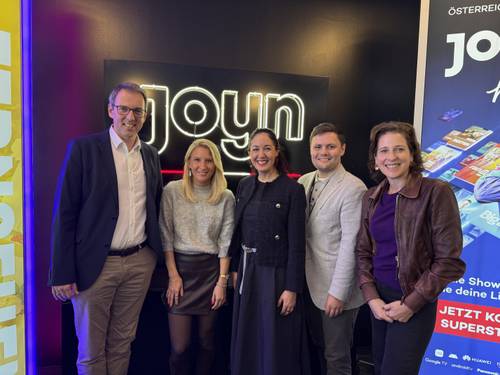 Fünf Menschen, die vor einem „joyn“-Neonschild lächeln, an dessen Seiten bunte Banner hängen.