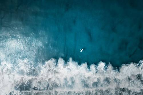 Imagine aeriană a unui surfer care vâslește pe o placă de surf în marea albastră, în timp ce valuri albe se sparg sub el.