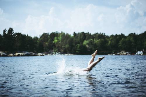 Person diving into a lake, legs visible above water, creating a splash; surrounded by trees under a partly cloudy sky.