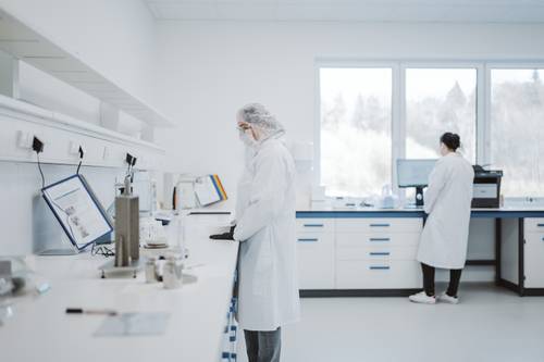 Două oameni de știință în halate albe de laborator lucrează într-un laborator luminos, cu echipamente și lumină solară pătrunzând prin ferestre mari.