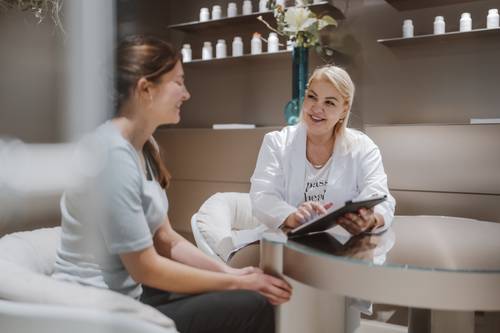 Eine Frau in einem weißen Kittel hält ein Tablet in der Hand und lächelt eine andere Frau an, die ihr gegenüber an einem Glastisch in einem modernen, gemütlichen Raum sitzt.