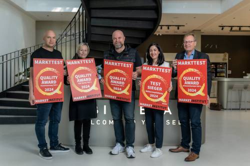 Fünf Personen mit Schildern „Markttest Quality Award 2024” und „Test Sieger” stehen vor einer Wendeltreppe.