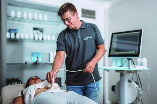Ein BIOGENA Biohacking & Diagnostics-Techniker überprüft die Knochendichte eines Patienten in einem modernen Behandlungsraum mit einem Computermonitor und Regalen voller BIOGENA-Produkte im Hintergrund.
