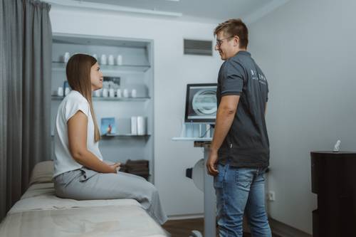 Eine Frau, die auf einem Liege sitzt, unterhält sich mit einem Mann, der neben ihr steht, in der Biogena Plaza; im Hintergrund ist ein Knochendichte-Messgerät zu sehen.