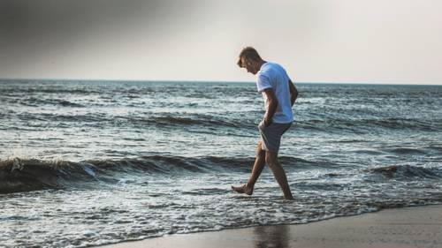 Ein Mann im weißen T-Shirt und Shorts geht am Strand entlang, während sanfte Wellen unter klarem Himmel an seine Füße spülen.