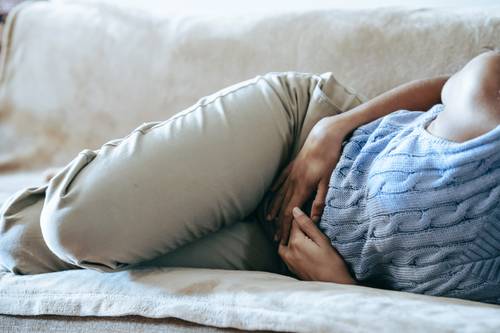 O persoană este întinsă pe o canapea, poartă un pulover albastru și pantaloni bej și pare să se țină de burtă de durere.