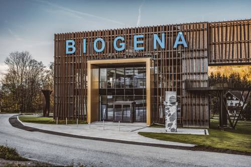 Ein modernes Gebäude mit der Aufschrift „BIOGENA“ an der Fassade, das sich durch eine Mischung aus Holzpaneelen und Glas auszeichnet und von viel Grün und einer geschwungenen Straße umgeben ist.