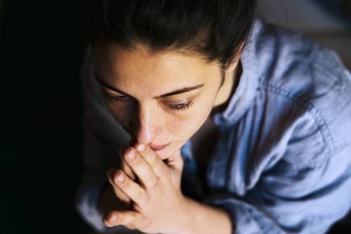 Frau in einem blauen Hemd, tief in Gedanken versunken, mit den Händen vor dem Gesicht verschränkt, von oben in einer schwach beleuchteten Umgebung betrachtet.