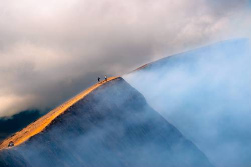Two hikers stand on a sunlit mountain ridge, surrounded by mist and clouds, highlighting a breathtaking landscape.