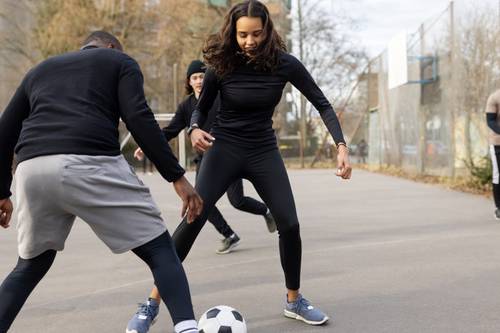 People playing soccer on an outdoor court, with a woman in black athleisure dribbling the ball and another person attempting to block her.