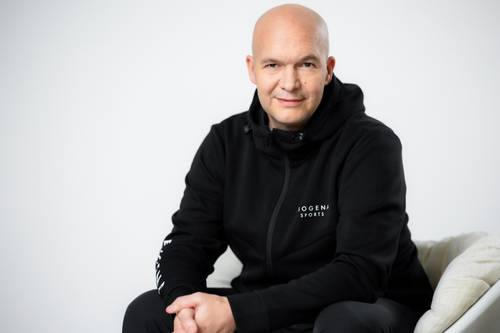 Ein glatzköpfiger Mann in einer schwarzen Jacke sitzt auf einem weißen Sofa, lächelt und hat die Hände gefaltet. Auf der Jacke ist das Logo „BIOGENA Sports“ zu sehen.