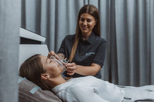 Eine Frau liegt entspannt auf einem Bett, während eine andere Frau ihr eine transparente Maske im Spa-ähnlichen Ambiente mit grauen Vorhängen aufträgt.