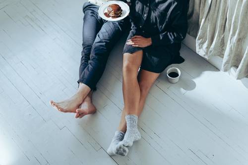 Two people are sitting on a white wooden floor, one holding a plate of pancakes. Next to them is a cup of coffee. Both are barefoot or wearing socks.